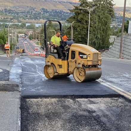 Central Paving crew member operating a roller over a fresh asphalt patch on a city street