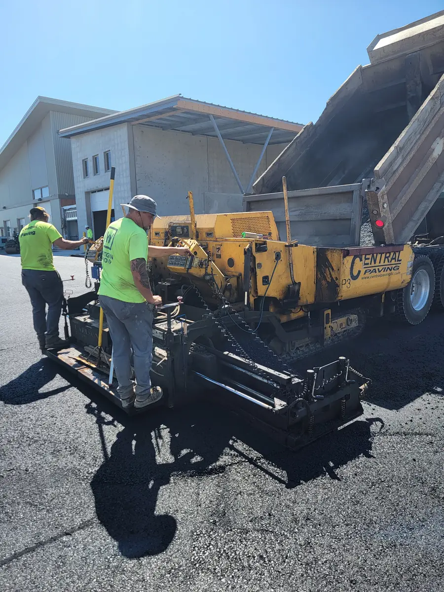 Crew members with a paver and dump truck in front of a building