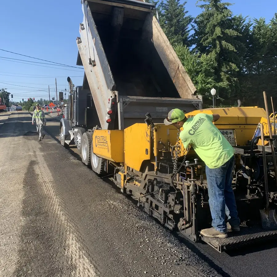 Dump truck feeding a yellow paver while a crew member works the screed