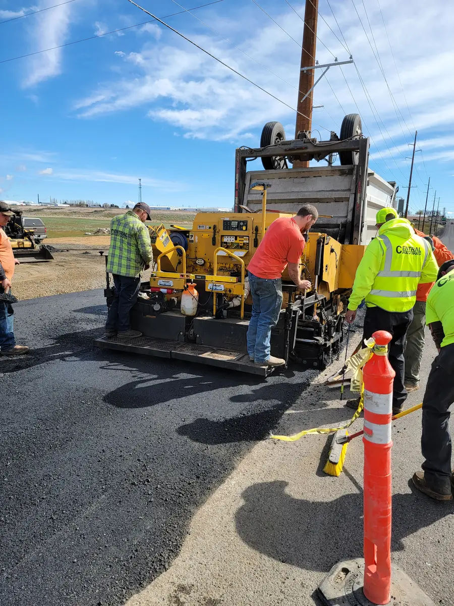 Paver and crew working on a road with an orange traffic cone in the foreground