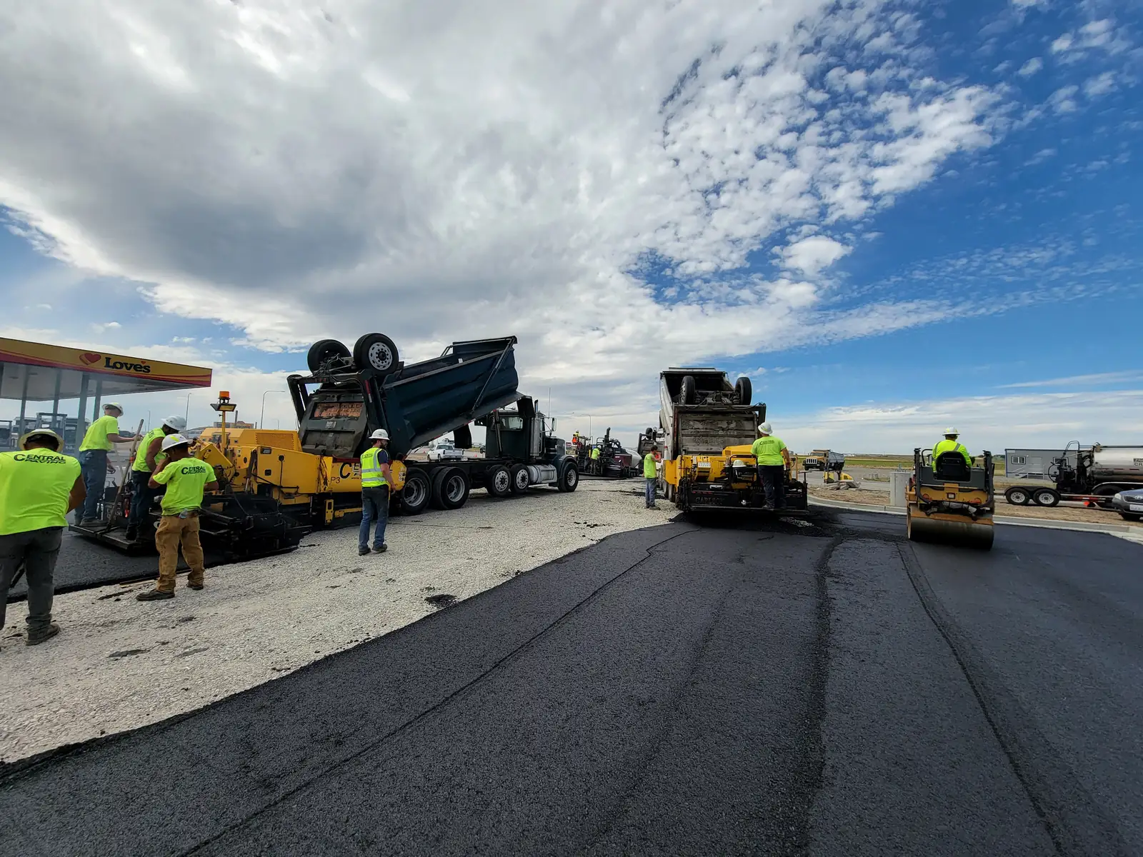 Dump truck, paver and rollers working on a lot beside a fuel station canopy
