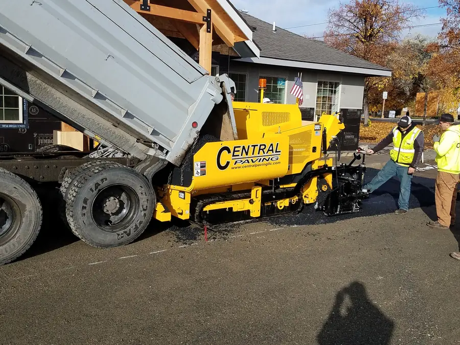 Dump truck feeding a Central Paving paver beside a log-style home