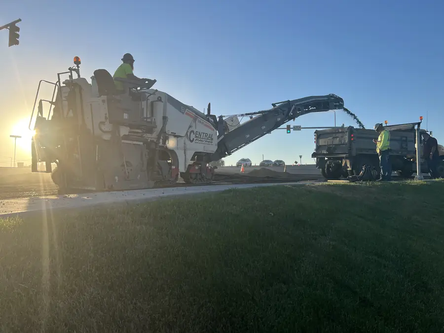 Milling machine loading a truck beside a lawn at sunrise