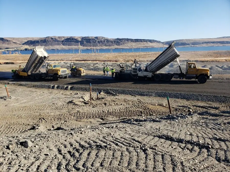 Dump trucks unloading on a graded gravel base beside a river with hills in the background